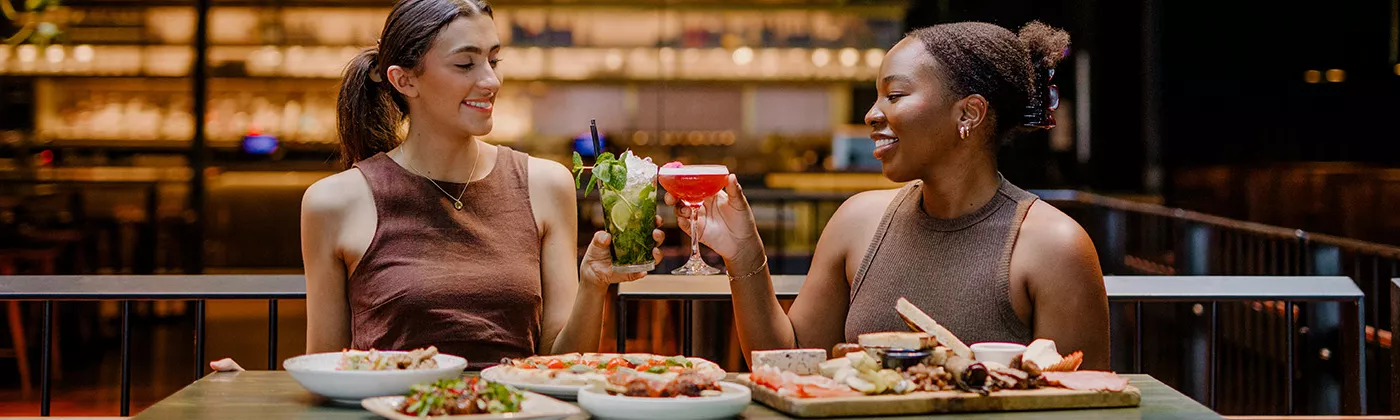 Two women in a restaurant with drinks in hand and meals on the table